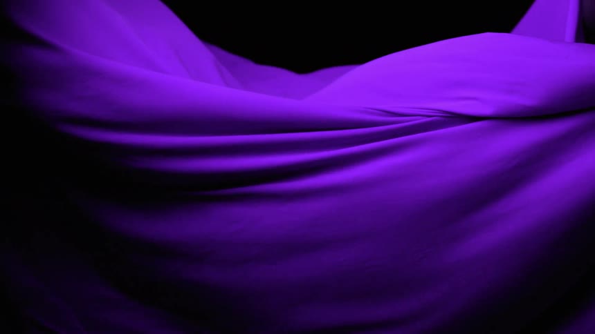 The video is a closeup shot of a large piece of smooth purple fabric moving slowly creating soft folds and wavesthe fabric is draped against a completely black background which makes the rich vibrant color and its subtle highlights stand out