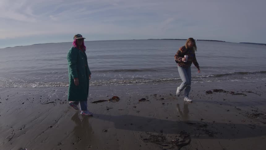 Two women walk in shallow water on a rocky beach under a cloudy sky
