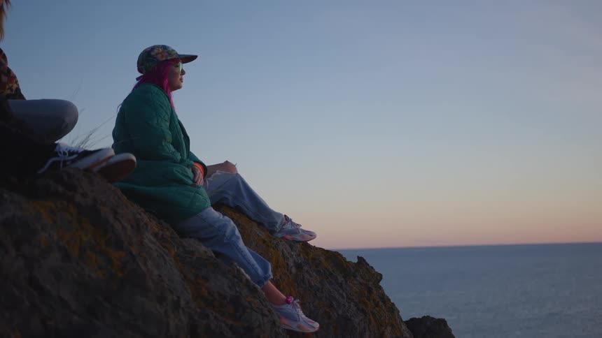 A side view of a young woman with pink hair wearing a baseball cap a green quilted jacket and jeans sits on a dark rocky cliff overlooking the ocean at sunset