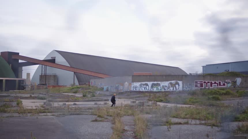 A long view of a dilapidated industrial landscape shows a man skateboarding in a dusty lot toward a large warehouse building with an elephant graffiti mural and a steep conveyor belt structure under a gray sky