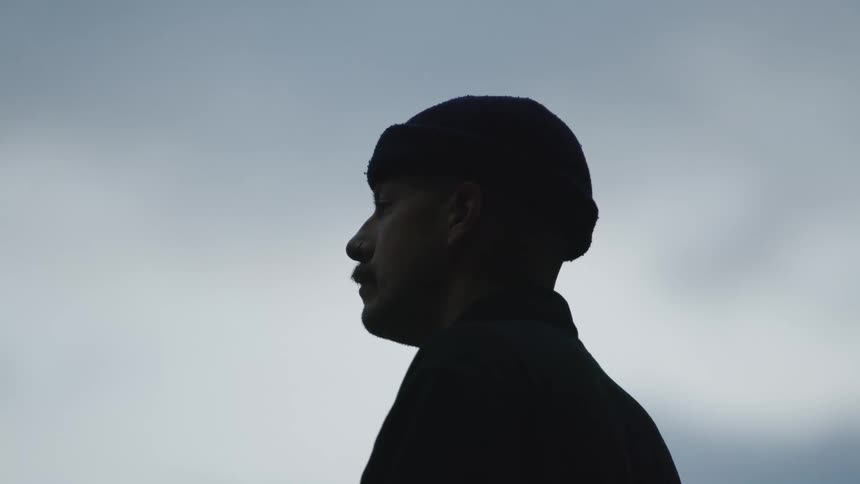 A man in profile wearing a beanie is silhouetted against an overcast sky