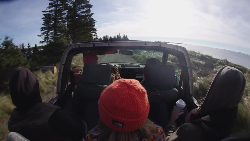 The video is a pov shot from the back seat of a vehicle with its top down driving along a rough road next to a large body of watertwo people are visible in the front seats and a person in the back with an orange knit hat is also visible all