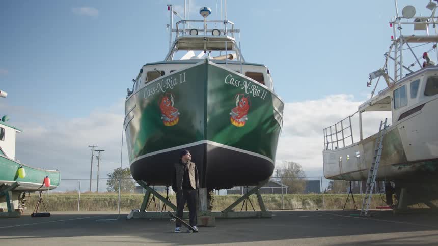 The man stands with his skateboard in front of a large boat in the lot