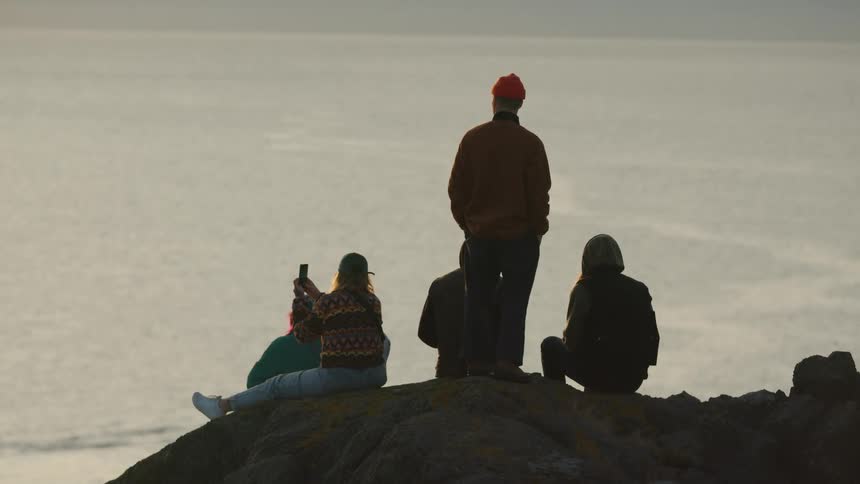 Five people stand and sit on a rocky hill overlooking an expanse of flat water one person stands and the others sits looking out one of the persons holds a phone up to take a picture against the hazy horizon