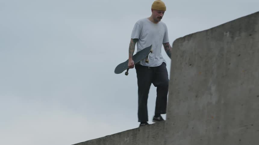 A man wearing a mustard beanie gray tshirt and dark pants walks along a concrete ledge while holding a skateboard