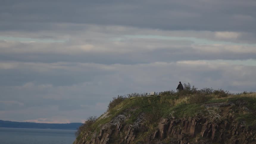 The video is a long shot of a lone figure walking along the top of a rugged coastal cliff or large grassy hillthe scene is dominated by a cloudy overcast sky and the dark rocky terrain conveying a sense of isolation or contemplation