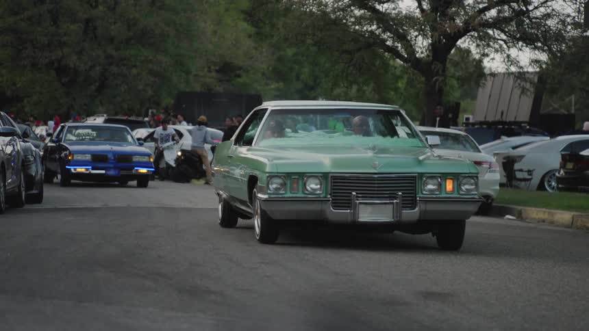 A vintage light green cadillac slowly cruises down a street crowded with other classic cars and people during an outdoor car meet or show