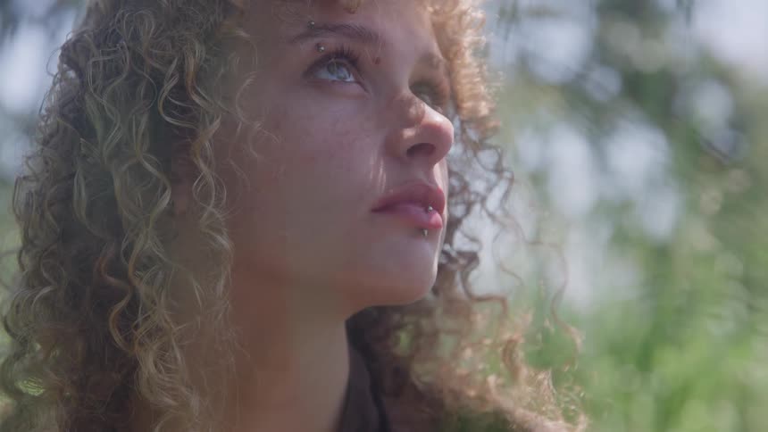 The video is a closeup shot of a young woman with light curly hair looking up and slightly to the sideshe has a lip ring and a piercing above her eye and the background is a soft blur of green foliage suggesting she is outdoors
