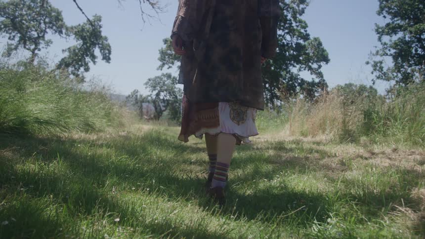 A person wearing a dark flowing top a patterned skirt striped socks and shoes walks across a grassy field on a bright sunny day