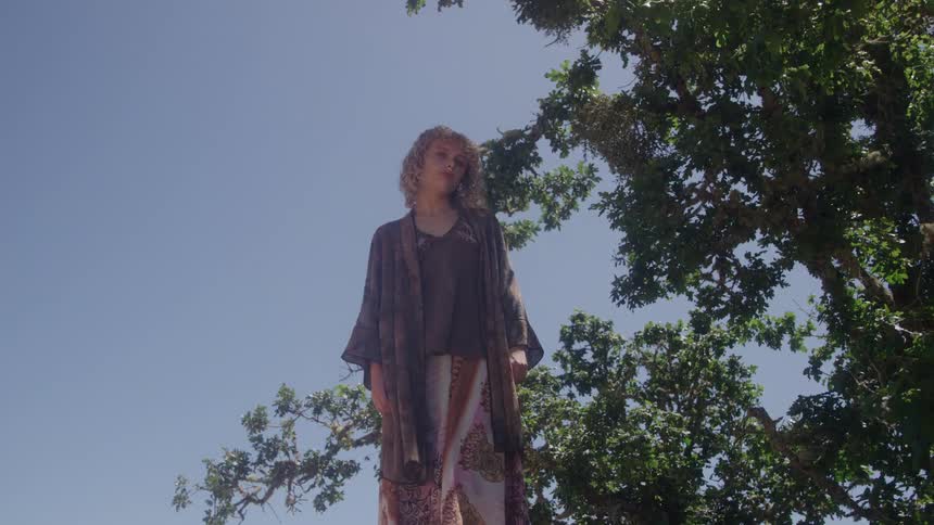 A blonde woman wearing a long dress is standing in a park next to a large tree