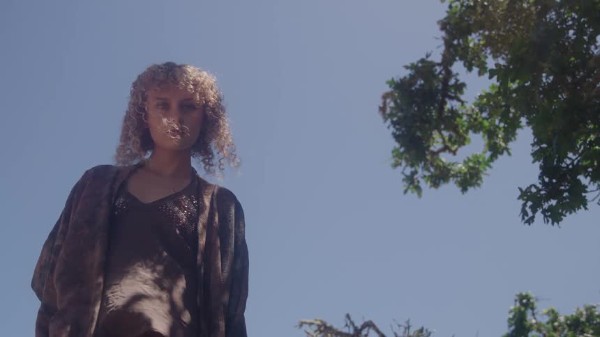A young woman with long curly hair stands beneath a tree on a sunny day
