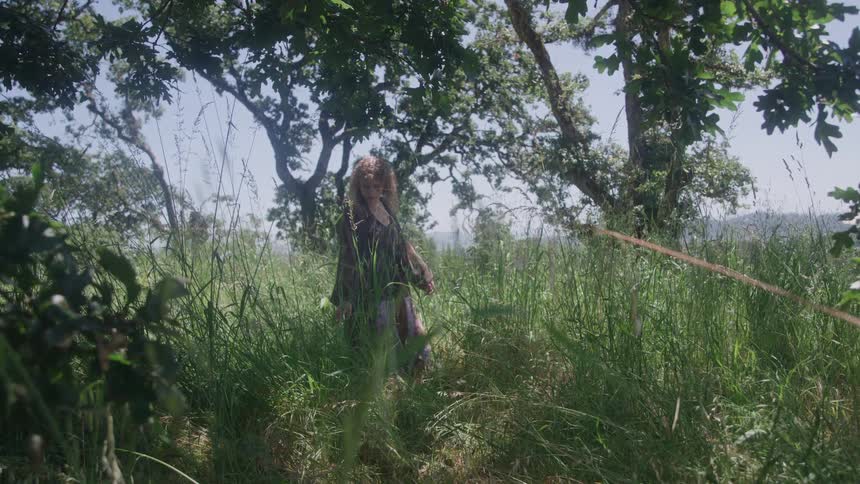 A young woman walks across a grassland surrounded by trees on a sunny day