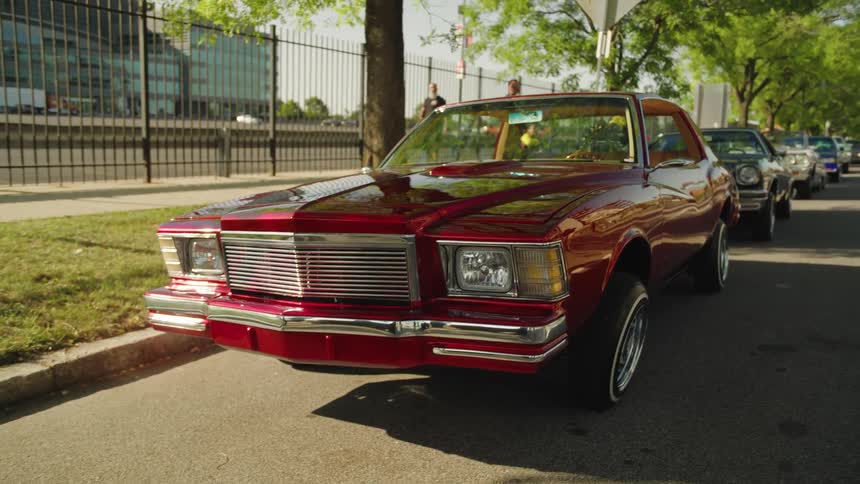 The video is an exterior lowangle shot of a classic shiny red car parked on a residential street during the daythe car which appears to be a chevrolet monte carlo or similar lowriderstyle vehicle is highly polished with chrome trim reflecti
