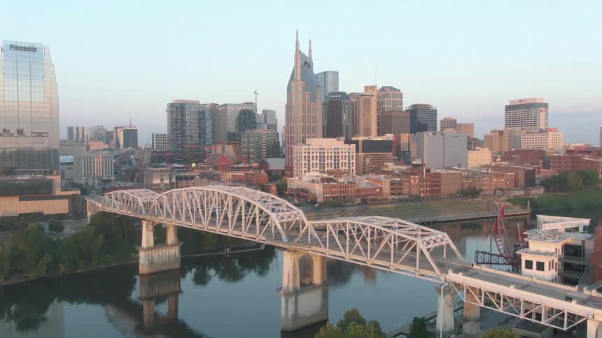 The video is an aerial closeup of a bridge spanning a river with a lush green park in the foregroundin the background the dense nashville tennessee skyline rises up dominated by modern highrise buildings including the distinct batman bu