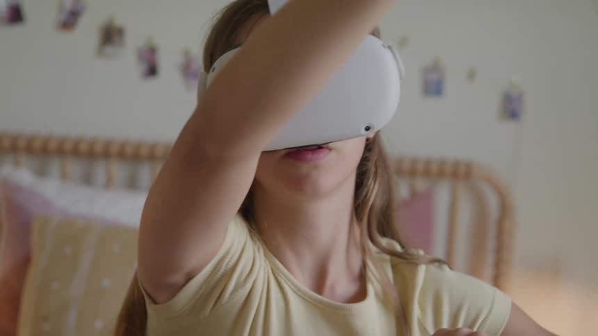 The video shows a young girl possibly a preteen with light brown hair wearing a virtual reality vr headset and a light yellow topshe is actively moving her arms as if interacting with a virtual environment with a blurry bedroom setting in