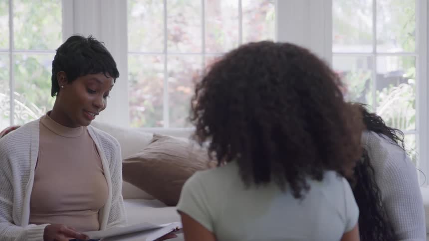 Three women sit together indoors near a wall of windows two of the women face the camera and speak or listen attentively one woman holds a book and the third woman faces away from the camera
