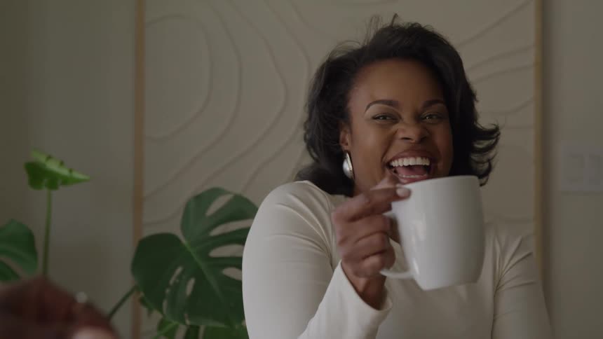 A woman with a joyful wide mouthed laugh holds a white mug and engages in conversation with a person in the foreground creating a warm and candid scene