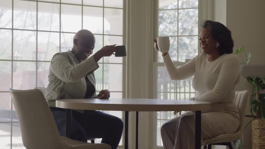 The video shows two person sitting across a small circular table from each other in a brightly lit roomthey both smile and raise their coffee mugs to touch in a friendly gesture suggesting a casual conversation or toast