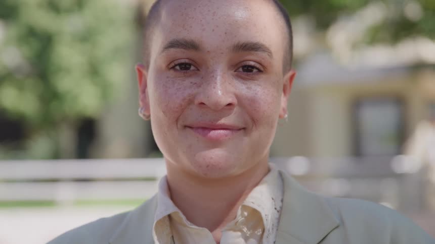 A woman with a shaved head smiles while posing outdoors