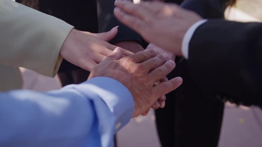 A close view shows diverse people in business attire stacking their hands together signifying teamwork and then pulling them away in a celebratory manner outdoors