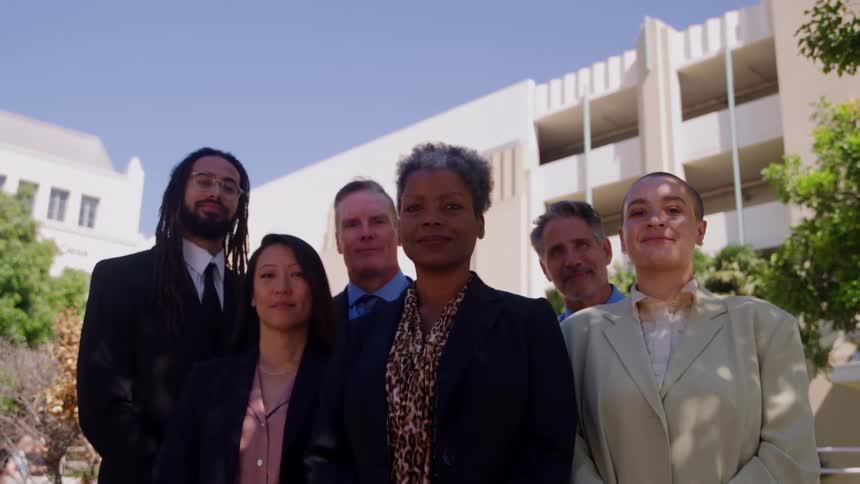 The video shows six people dressed in business attire standing together outdoors they appear to be a diverse group of colleagues or professionalsthey are standing in front of a white building complex under a clear sunny sky with some green trees a
