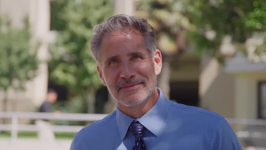 A middleaged man with gray hair and a blue collared shirt with a dark necktie stands outdoors looks directly at the camera and smiles slightly against a softfocus backdrop of green trees and white railings