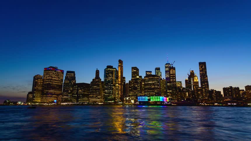 A panoramic view of the brilliantly lit new york city skyline with many tall skyscrapers is reflected on the surface of the dark blue water at twilight