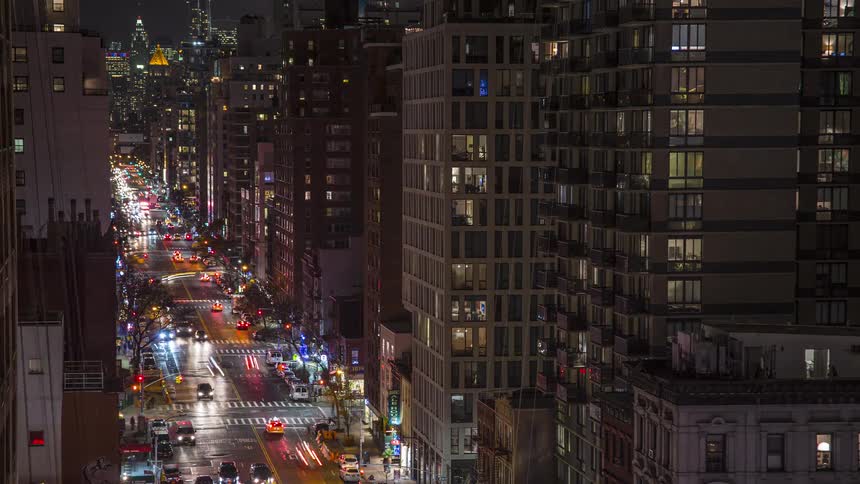 A street flanked by tall buildings is illuminated at night showing moving traffic blurring into light streaks while the numerous windows of the surrounding residential structures glow with interior illumination