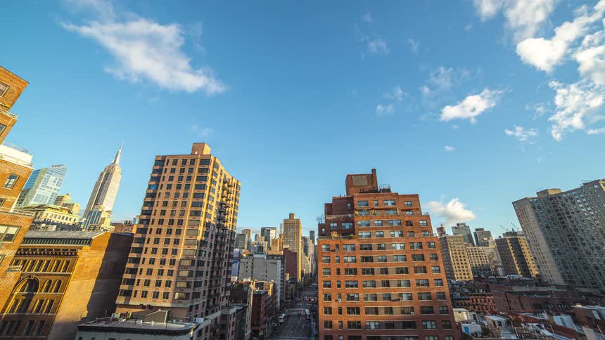 A wide view of a crowded city shows numerous tall rectangular buildings under a clear blue sky with white clouds the scene is on a time lapse