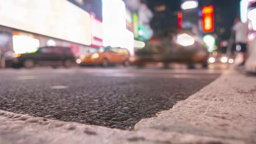 This is an extreme lowangle shot of a city street at night focused sharply on the textured asphalt road and the concrete curb in the foreground in the blurry background cars and trucks drive past as the street is lit by the intense outoffoc