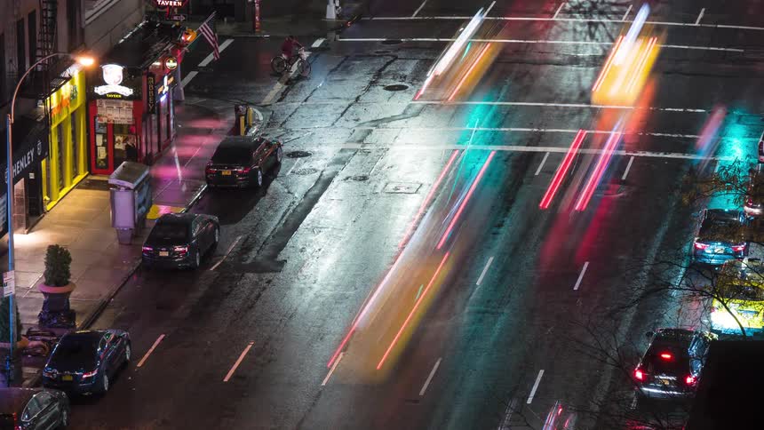 The video is an elevated wide shot of a wet city street at night with vehicles driving on itthe main focus is the motion blur of the cars taillights and streetlights streaking in red and white across the dark reflective asphalt