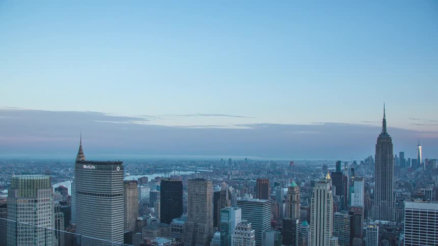 The video provides an elevated view of a dense cityscape against a bright blue sky likely new york citythe famous empire state building is prominent on the right surrounded by many other highrise buildings that stretch toward the horizon