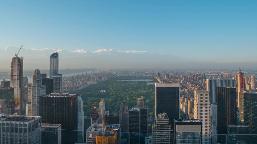 A camera captures a dense collection of tall rectangular buildings surrounding a large central green area under a bright hazy sky emphasizing the contrast between the structures and the park