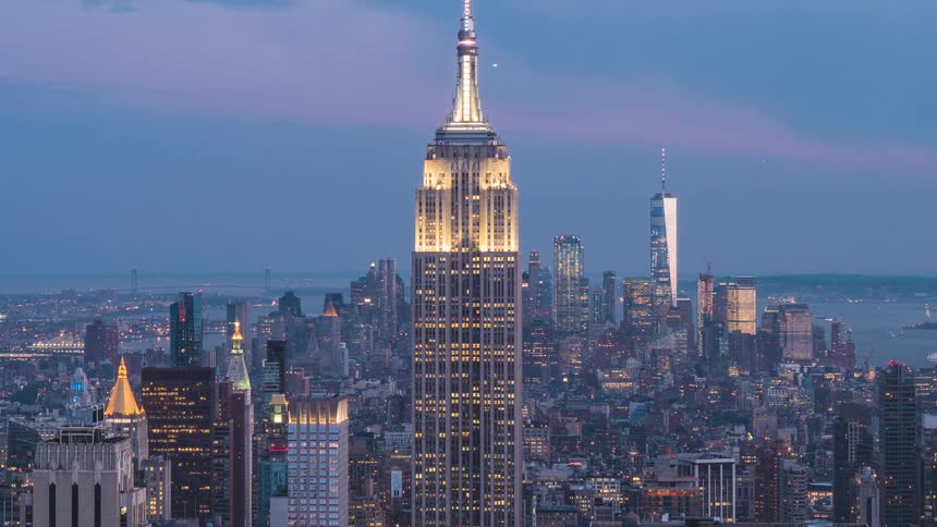 A central tall skyscraper is brightly illuminated against a fading purple and blue sky standing above a dense urban area where numerous buildings starts to glow in a time lapse scene