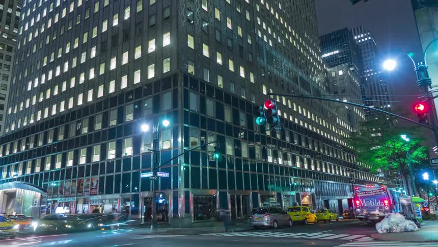 A time lapse shows traffic including yellow taxi cabs moving quickly past a large modern skyscraper with many illuminated windows in a busy city intersection at night