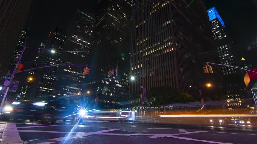 This is a timelapse shot of a busy city street corner at night showcasing a large modern intersection surrounded by towering illuminated skyscrapers the blurred trails of car headlights and taillights streak across the wet pavement emph