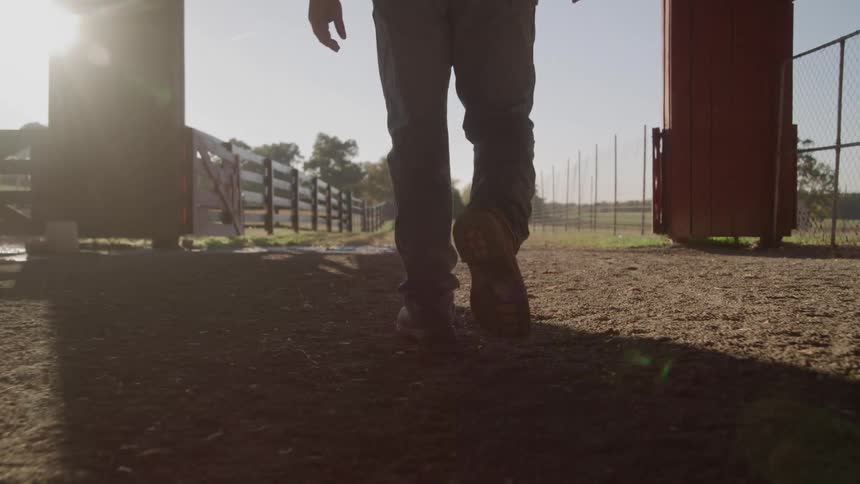 The video is a lowangle shot of a mans legs and feet as he walks away on a dirt path under the cover of a barn or shedsunlight streams in from the open end of the structure casting long shadows of the man and the wooden posts onto the ground