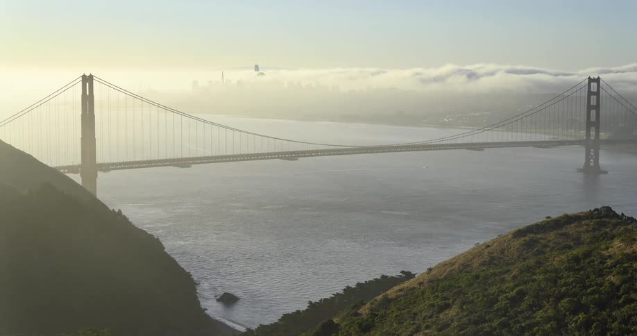 A large redorange suspension bridge spans a body of water between two green hills while thick lowlying clouds obscure a distant urban landscape under a sky with sun haze