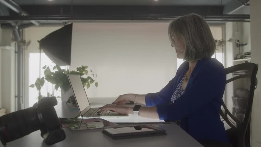A professional gray haired woman wearing a blue blazer sits at a desk in a bright photography studio typing on a laptop with a camera in the foreground and studio lighting in the background