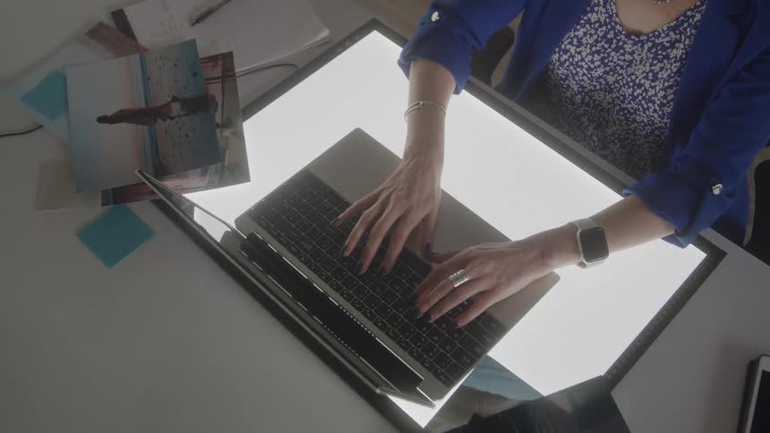 Woman sitting at a desk typing on a laptop computer as she reaches for a pen