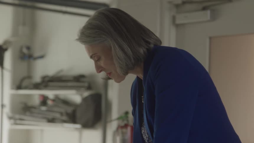 A middle aged woman with gray hair wearing a blue blazer works on a brightly lit drafting table with paper and tools in a design or engineering office