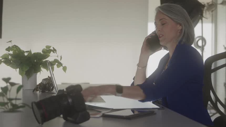 A woman photographer answers her smartphone while sitting at the desk and working on her laptop