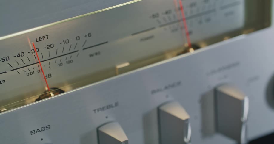 A closeup shot shows the silver faceplate of an audio component featuring a left channel volume meter with a red needle and printed decibel scale and partially visible control knobs