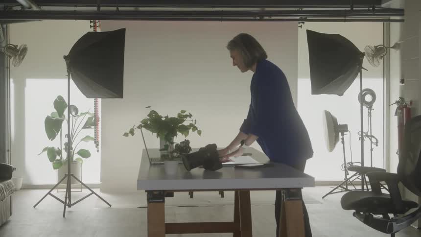 A woman in a blue jacket stands at a white desk in a studio setting writing on a piece of paper next to a laptop camera and plant while studio lights on stands frame the scene