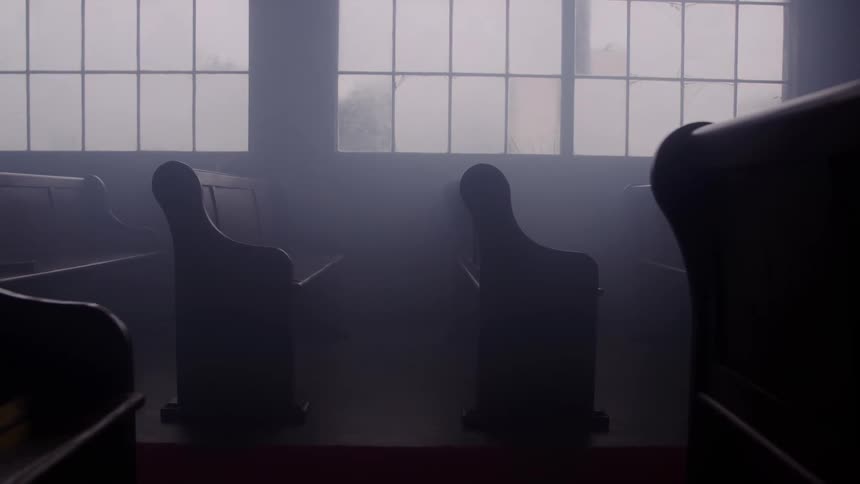 A view from a shadowy dark church aisle shows the silhouettes of wooden pews facing large windows that look out into thick mysterious fog