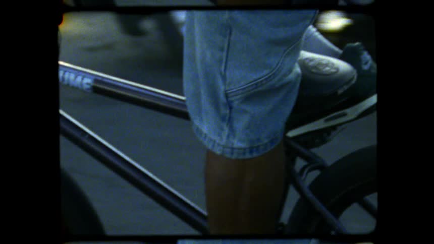 A close angled shot with an old film border shows a person wearing light blue jeans placing their dark shoe onto the pedal of a black volume bmx bike