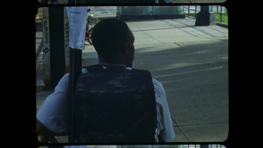 A low angle dark rear view shows a man wearing a white shirt and a dark vest sitting in a wheelchair on a sunny city sidewalk next to a sign pole