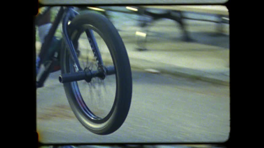 A person in a white shirt and shorts rides a black bmx bicycle on a paved path performing a wheelie with the front wheel lifted off the ground