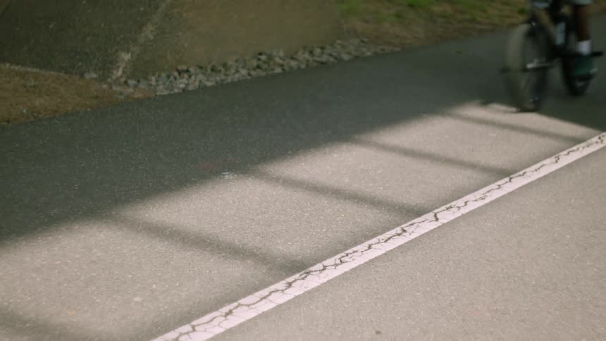 The video is a lowangle closeup shot of a person riding a bicycle on a dark paved path next to a cracked white linethe riders feet and the bicycles wheel pass quickly over the pavement partially obscured by a dark shadow on the left side of