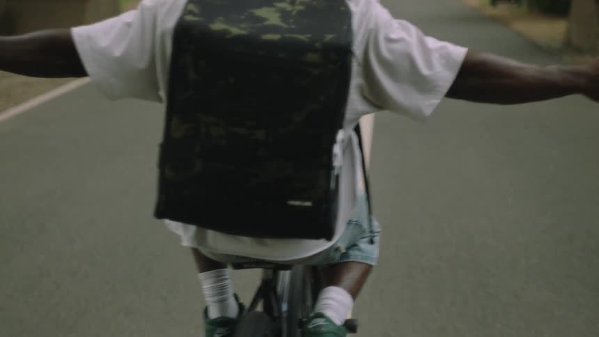 A close view from behind shows a man wearing a white shirt and a camouflage backpack riding a bicycle down a tree lined path with his arms spread wide in a gesture of freedom